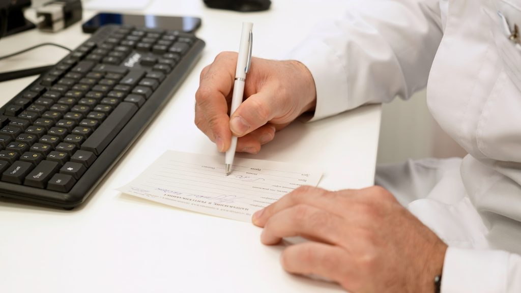 Hands of male doctor writing prescription on paper. Clip. Desk of a doctor with computer keyboard